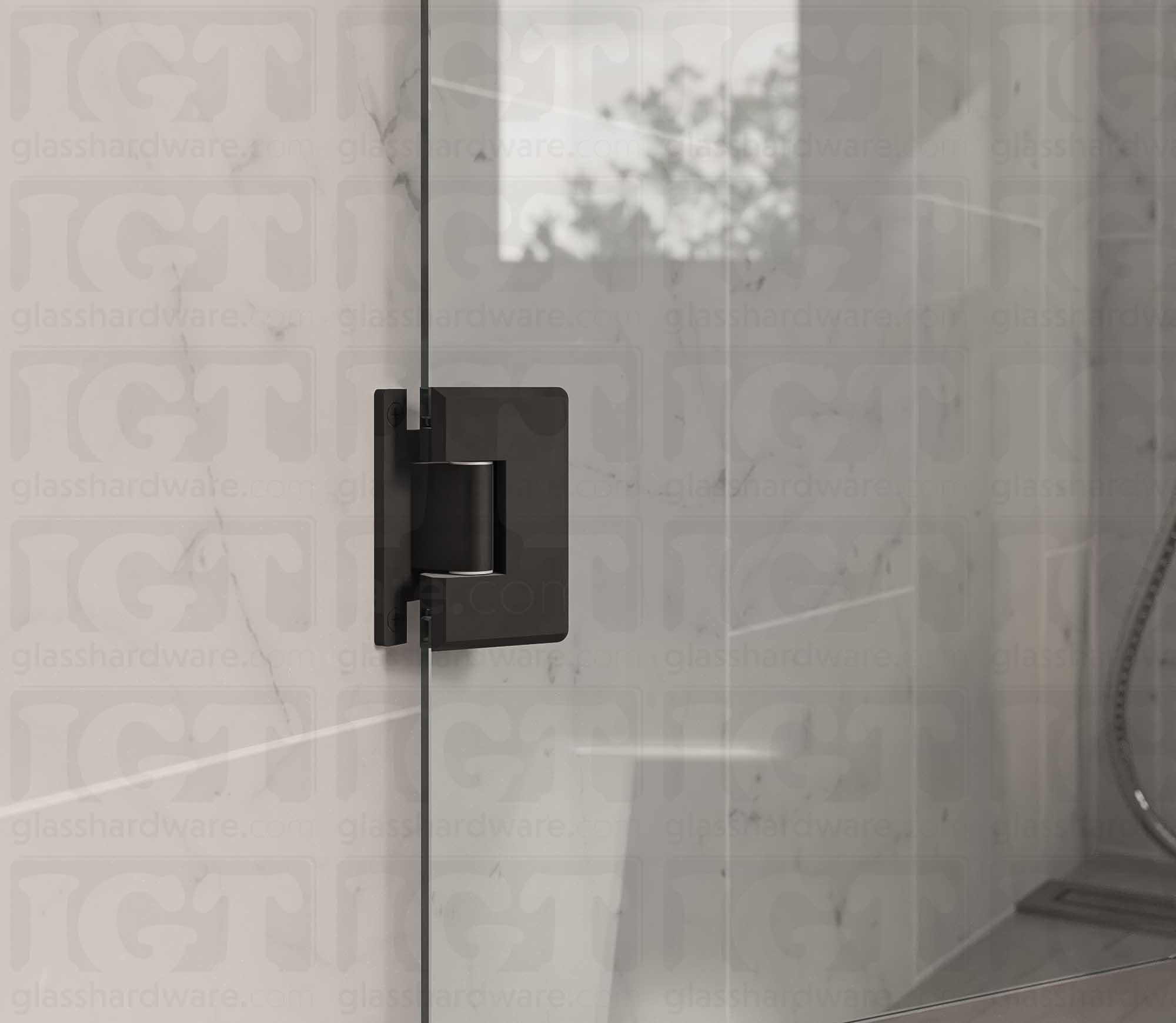 A close up view of the Heavy Duty Wall-to-Glass “H” Back Bilboa Hinge installed on a frameless glass shower door. The bathroom's natural lighting reflects along the hinge's sleek beveled edges, adding dimension to the overall design. Matte Black.