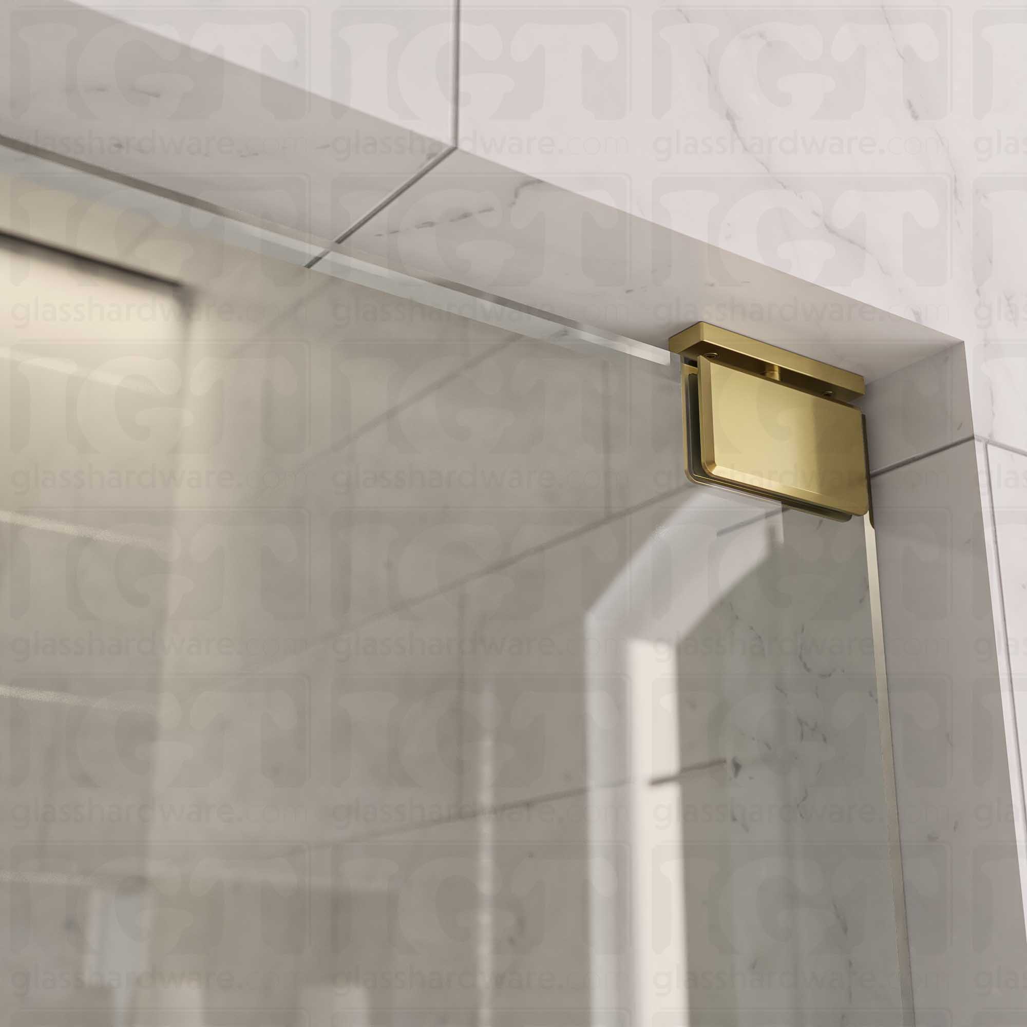 Close-up of the Top and Bottom Bilboa Pivot Hinge mounted at the top of the glass shower door, securing it to the tiled wall. The hinge has soft rounded corners and beveled edges, making it blend into the elegant bathroom setting. Gold Brushed.